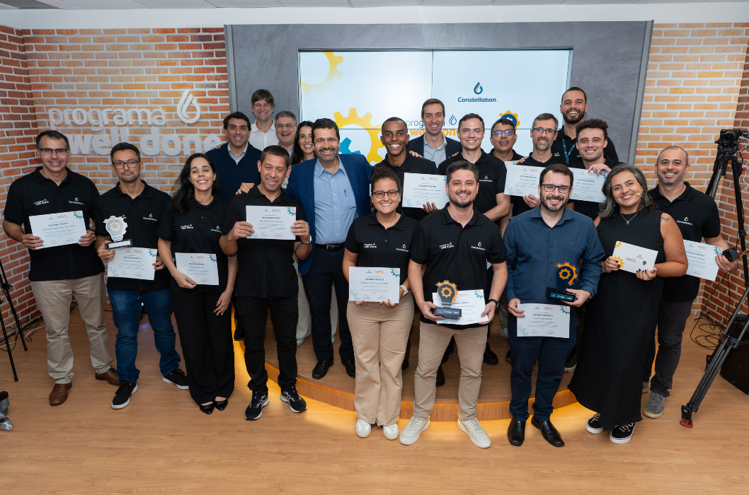 Group photo of an awarded Constellation team. About 15 employees, men and women, pose smiling in front of a brick wall with the 'well done program' logo. Several of them hold white paper certificates and small trophies with gear icons. The atmosphere is bright and festive.