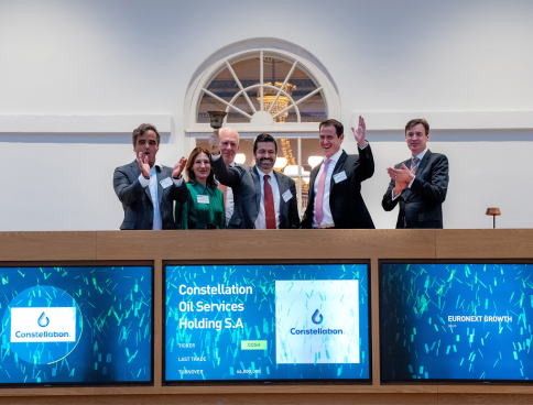 Group of six executives in formal attire on a ceremonial balcony, applauding and smiling. Below them, blue digital panels display the Constellation logo and the text 'Constellation Oil Services Holding S.A', celebrating the stock exchange listing.