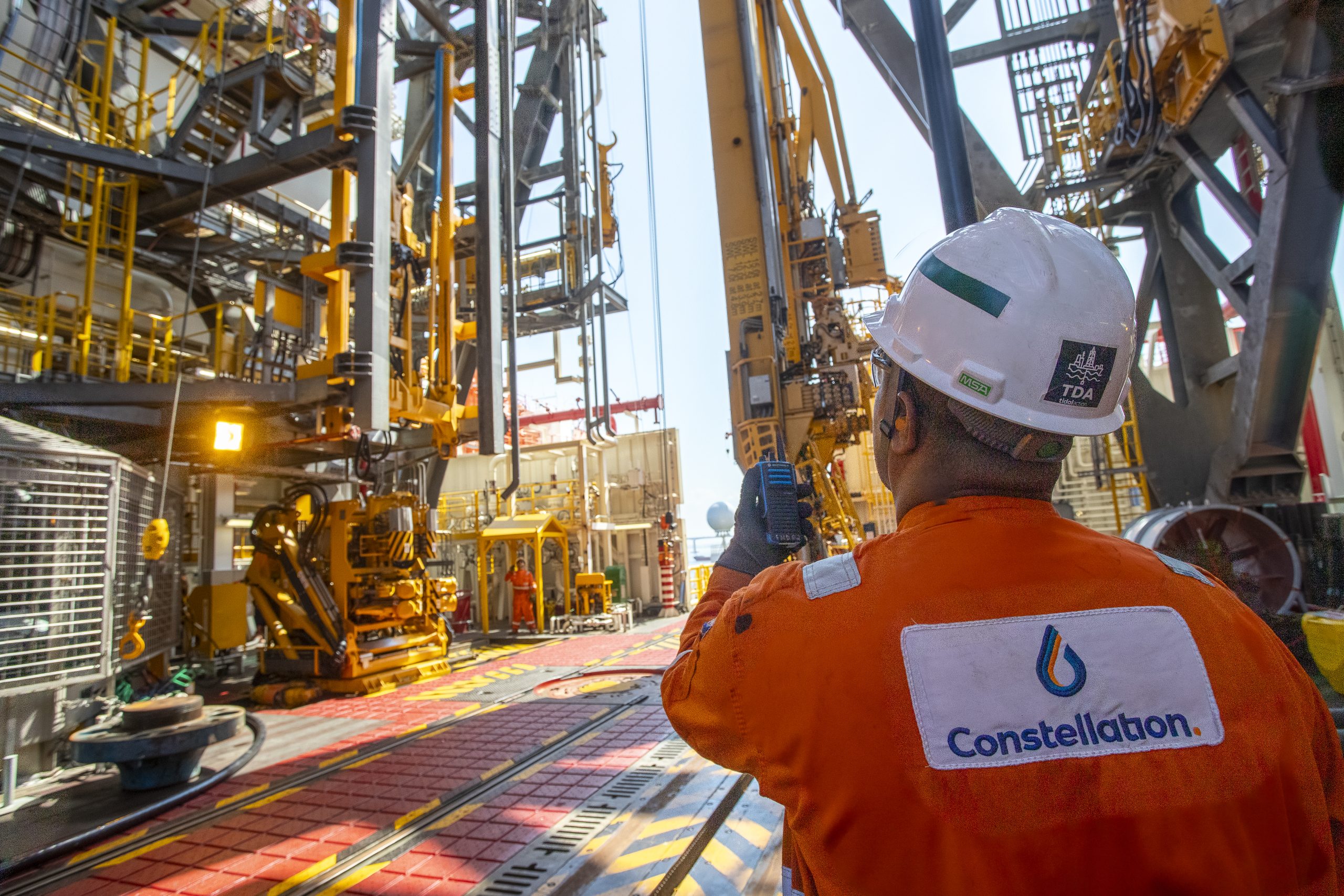 Constellation worker in orange overalls and white helmet, seen from behind, looking up toward the base of the derrick and complex equipment on a rig deck.