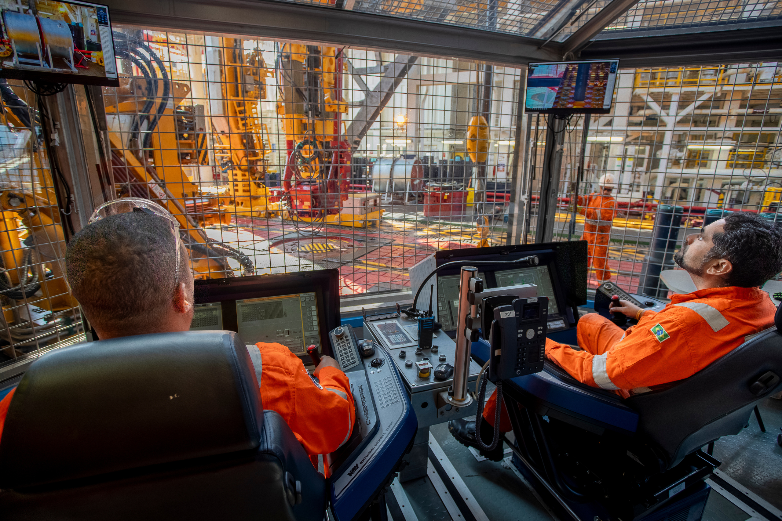 Operator sitting in a modern and high-tech drilling control cabin (driller's cabin), surrounded by several computer screens displaying graphic data and holding ergonomic command joysticks.
