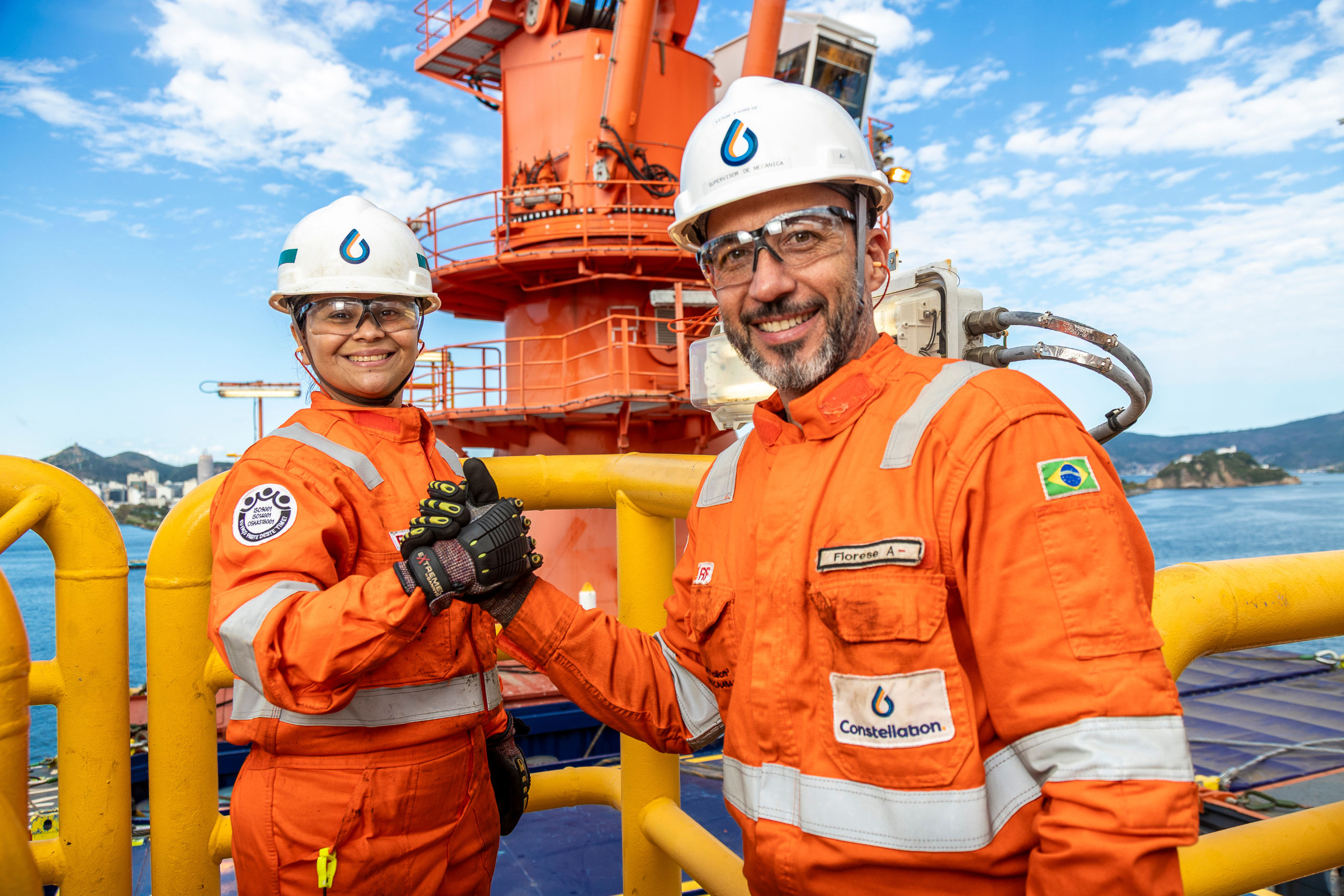 Photo of two Constellation employees, a man and a woman, wearing orange overalls, white helmets and safety glasses. They are on a sunny offshore platform, shaking hands and smiling directly at the camera. In the background, yellow metallic structures, the sea and a blue sky with clouds are seen.