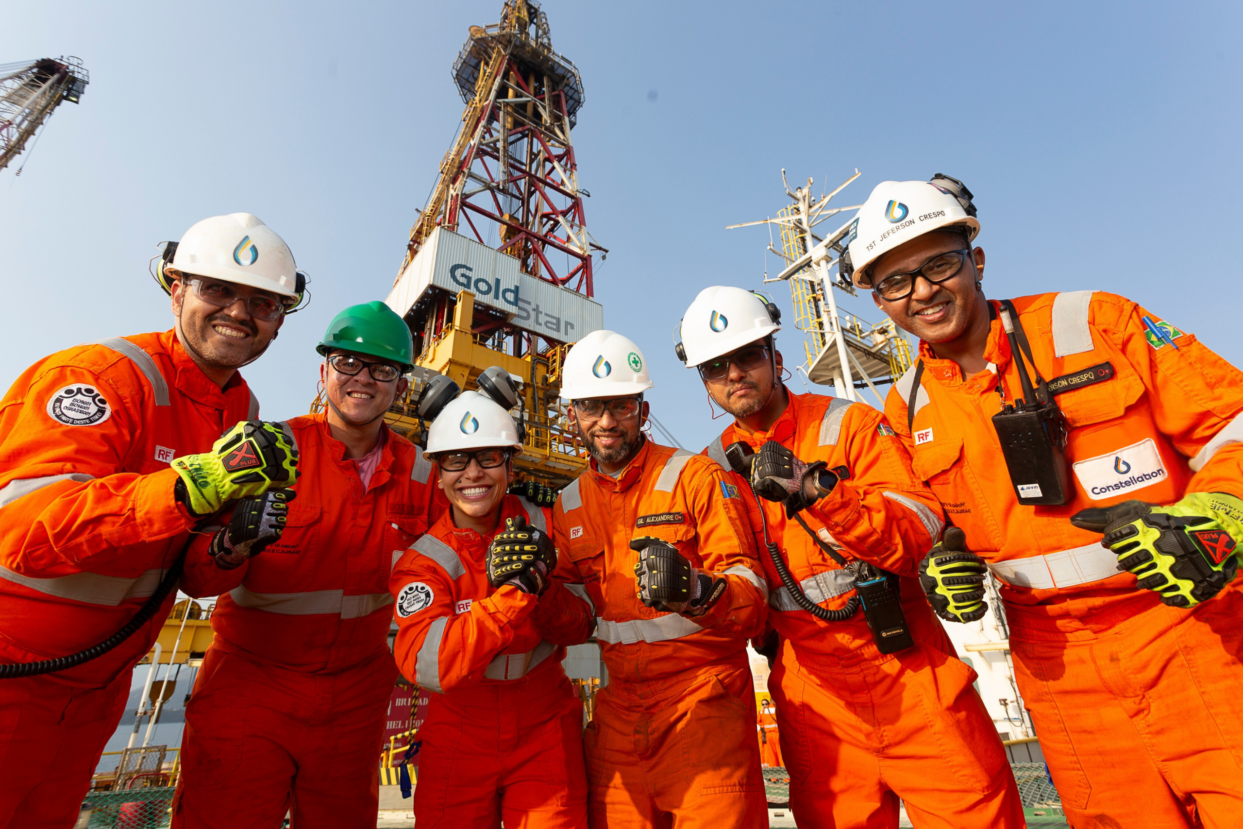 Photo of an excited group of six offshore workers, men and women of different ethnicities, embracing and smiling, posing for the camera. They wear orange overalls and white helmets. In the background, the derrick of the platform is seen with the name 'Gold Star' written.