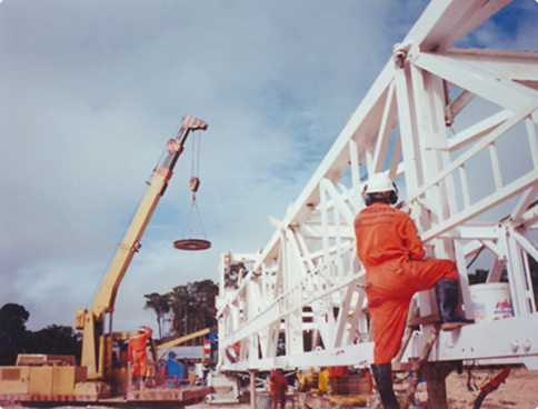 Color historical photograph showing onshore operations. A white latticed drilling structure is seen being hoisted or assembled by a yellow crane, with an orange overall worker observing the operation with their back to the camera under a cloudy sky.
