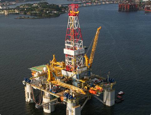 Aerial view of a drilling platform anchored in a calm water bay, near an urban coastline with bridges and buildings in the background. The platform has yellow cranes and a red and white derrick.