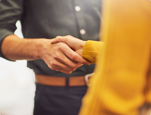 Close-up of a handshake between two people in a corporate environment. One person wears a dark gray dress shirt and the other a yellow piece of clothing. The image is blurred in the background, highlighting the gesture of agreement.