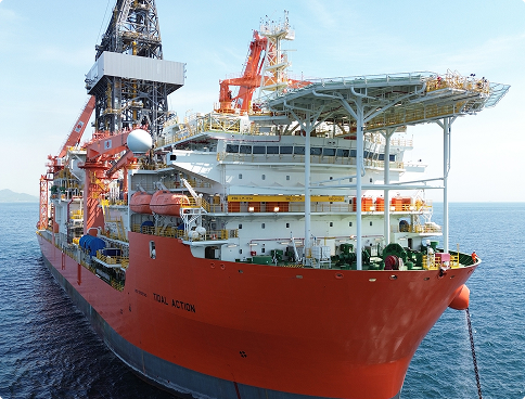 Angled front view of the 'Tidal Action' drillship. The hull is vibrant red and the superstructure is white, with a grated helideck projecting ahead of the bow. The vessel's name is visible on the hull in black.