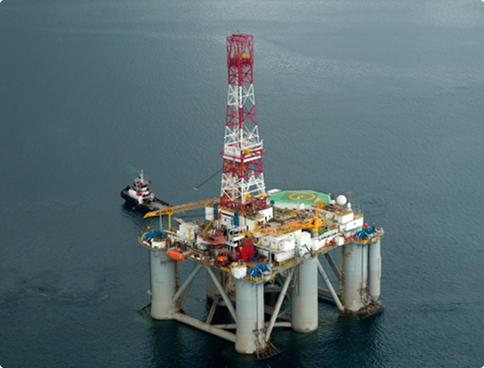 Aerial view of the 'Alaskan Star' semi-submersible platform in the open sea. The structure has large cylindrical white columns and a latticed derrick in red and white colors. A small black tugboat assists the operation near one of the columns.