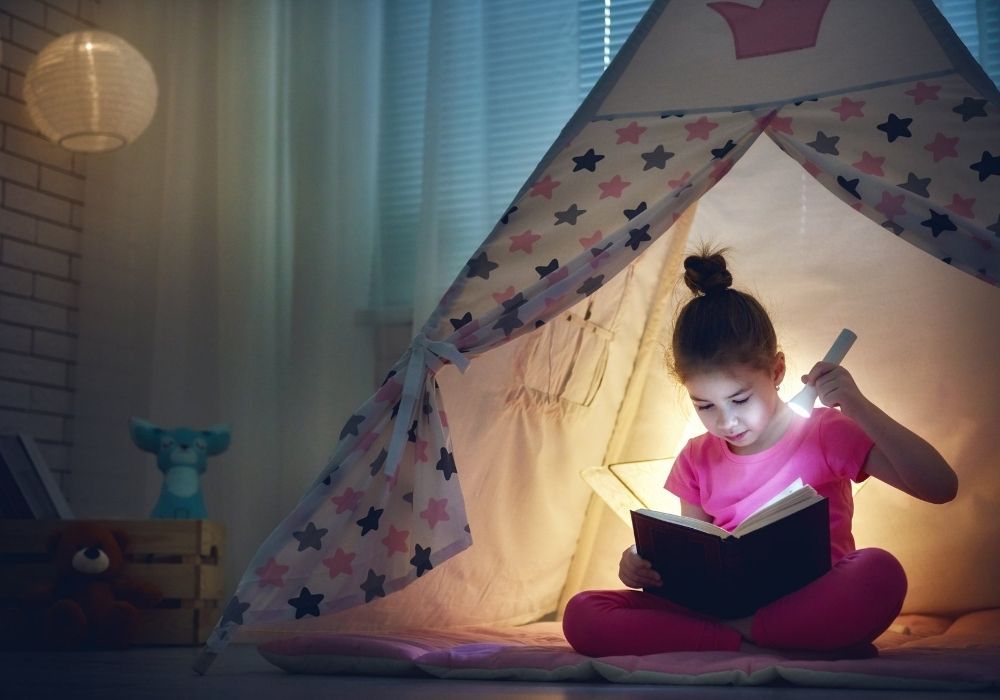 A girl in a play tent reading a book with a flashlight.