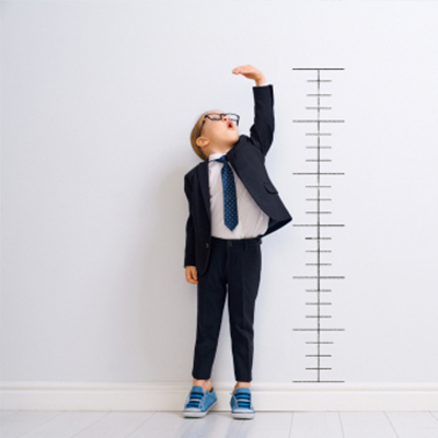 A young child in a suit stands against a wall, reaching upward toward a vertical height measurement scale.