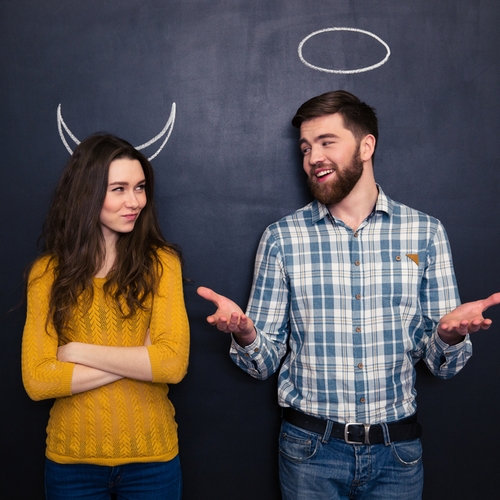 Smiling young man and woman imitating devil and angel standing over chalkboard background