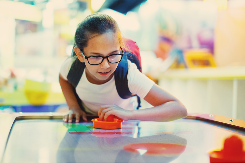 Young girl with glasses and wearing a backpack playing air hockey.