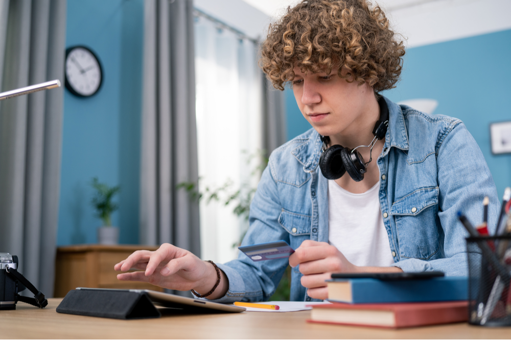 Concentrated millennial boy using tablet to pay with plastic credit card.