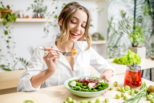 Young woman eating healthy food sitting in the beautiful interior with green flowers on the background.