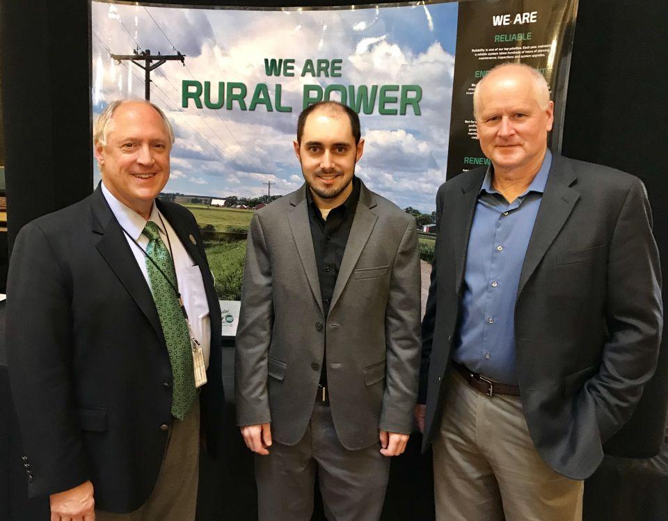 Two men from North West REC stand with Iowa Representative Tom Jeneary in front of a background image that reads: "We are rural power"