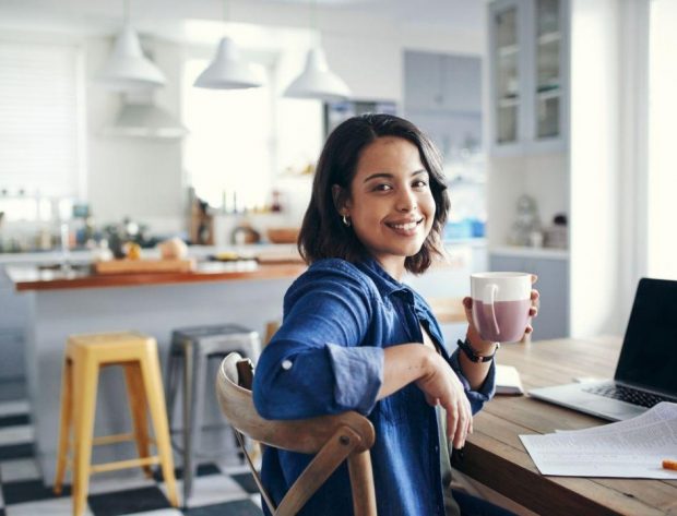 Photo of young woman working at home and sitting by her desk and computer. She is holding a coffee cup.