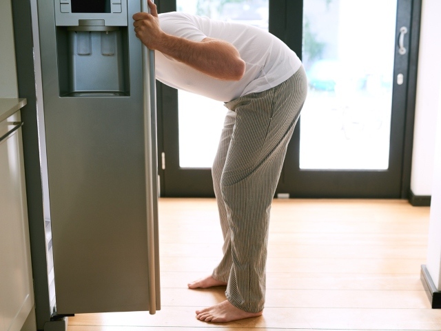 Photo image of a man in his pajamas peering into a refrigerator. The door is open.