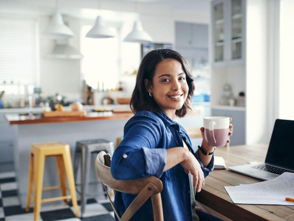 Photo of young woman working at home and sitting by her desk and computer. She is holding a coffee cup.