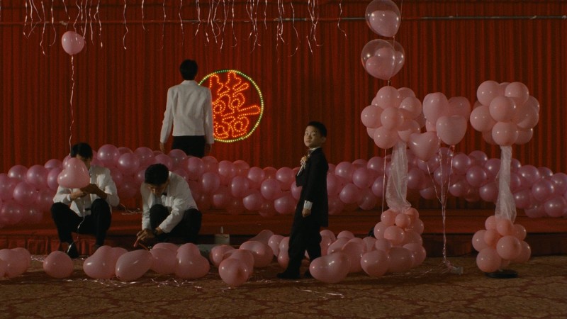 컬러 원경. 붉은 커튼과 분홍 풍선 장식 앞에 여러 사람이 흩어져 있다. 서 있는 사람과 낮게 앉은 사람들이 함께 보인다.