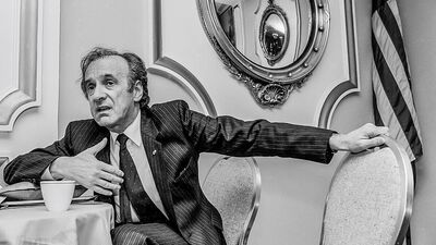 A person in a suit sits at a table, gesturing with one hand. An American flag and mirror are in the background.