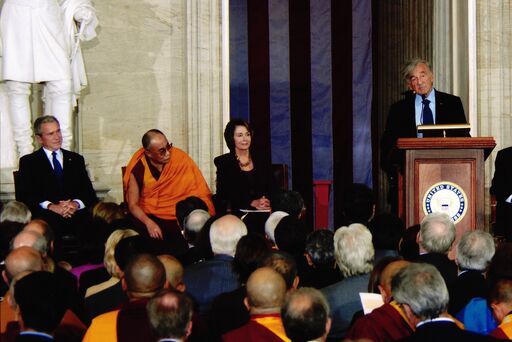 A speaker at a podium addresses an audience; officials and the Dalai Lama are seated on stage.