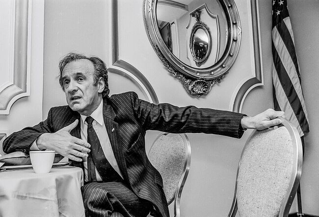 A person in a suit sits at a table, gesturing with one hand. An American flag and mirror are in the background.