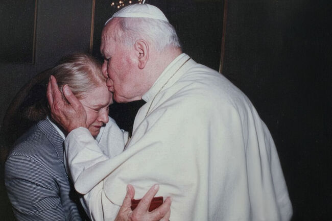 An elderly man in white religious attire kisses and comforts a person with blonde hair and a plaid jacket.