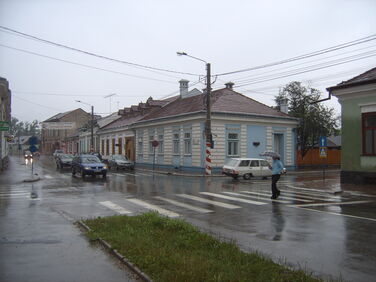 A one story house with blue shutters and trim shown on a rainy day with cars passing by on the street.