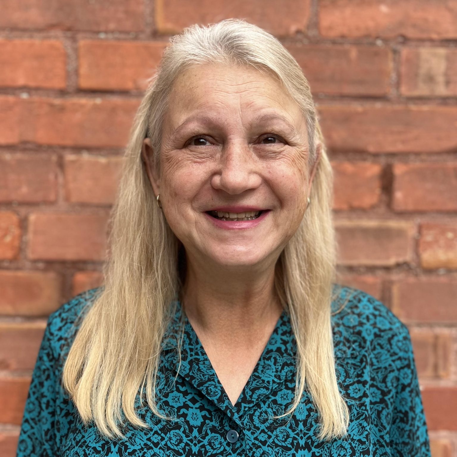 A person with long blonde hair in front of a red brick wall, wearing a teal floral-patterned shirt.