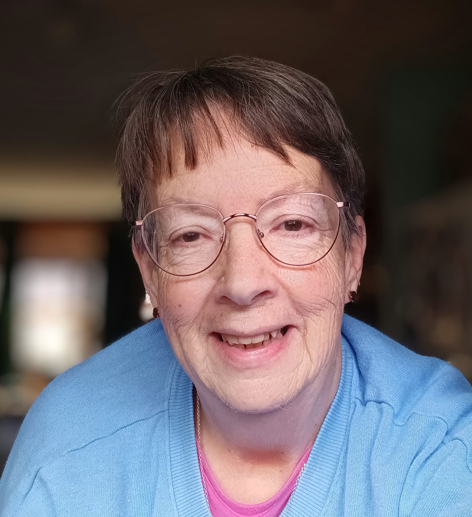 A woman with short hair and glasses smiles warmly. She wears a blue sweater over a pink top