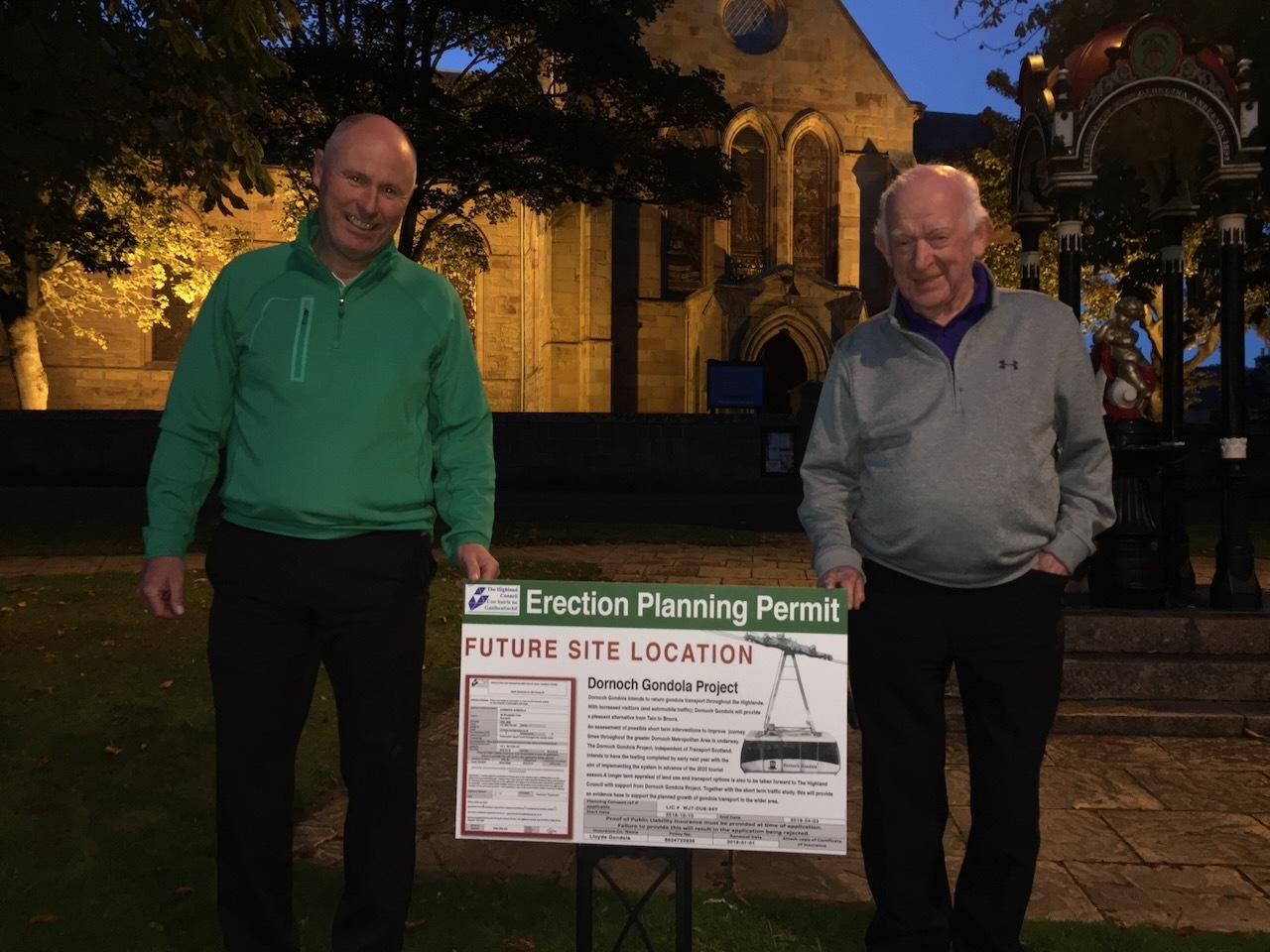 Two company representatives outside Dornoch Cathedral at night, holding the planning notice that marks the future site of Dornoch Cathedral Station.