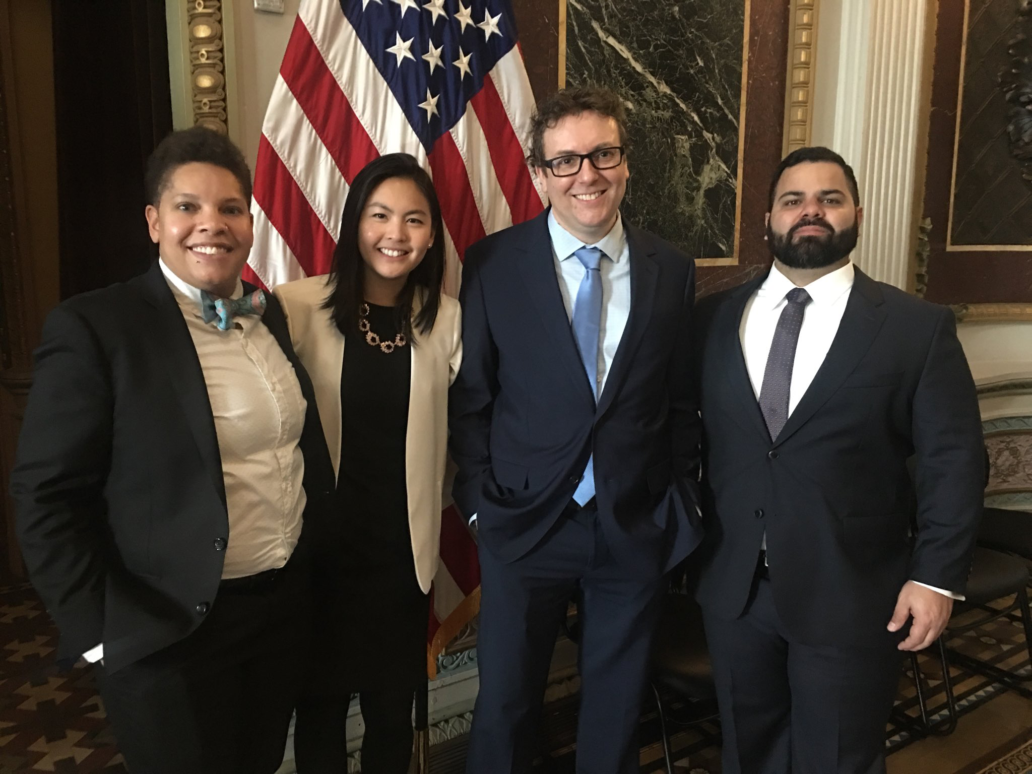 Clarice stands in front of a United States flag with three other Presidential Innovation Fellows. All are smiling and wearing professional-formal attire.