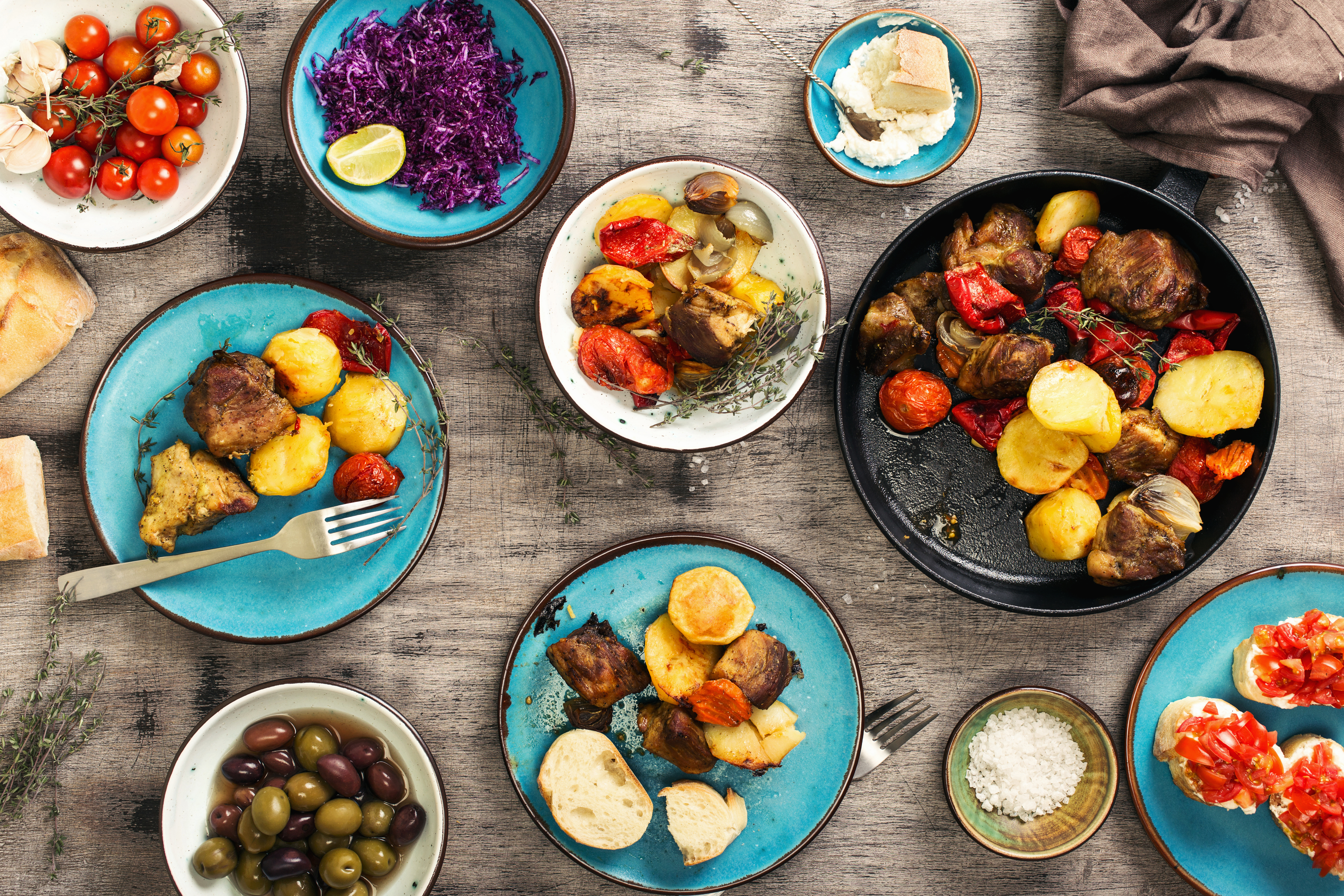 A photograph of a dinner table with plates, bowls and utensils. The dishes are holding olives, salt, bruschetta, tomatoes, cabbage, and bread, and there is a frying pan with potatoes and meat.