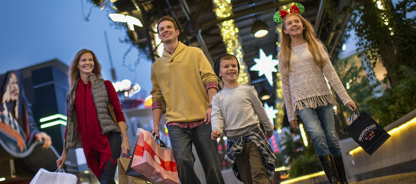 A family of four carries shopping bags and strolls underneath a decorated Sunshine Highline in the West Side of Disney Springs