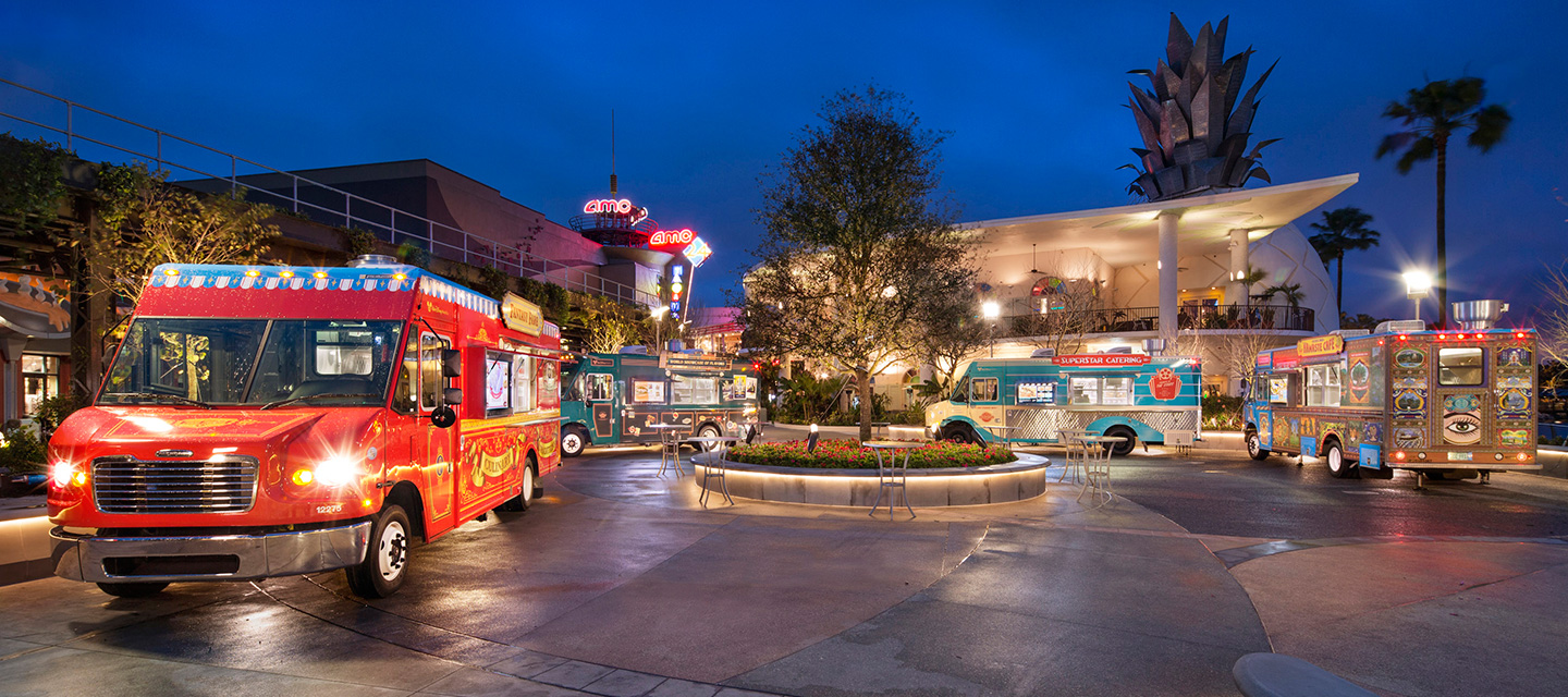 Four food trucks parked curbside within Exposition Park at dusk