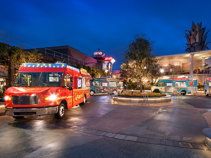 Four food trucks parked curbside within Exposition Park at dusk