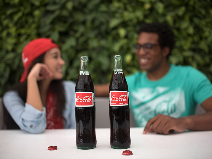 Two Coca Cola bottles with the caps off, rest on a table in front of 2 friends
