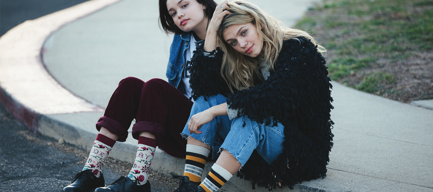 Two young females sit on a curb wearing stylish outfits and socks from the Stance sock boutique at Disney Springs