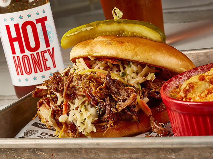A tray with a pulled pork sandwich with a pickle on top adjacent to a mini casserole dish of mac and cheese