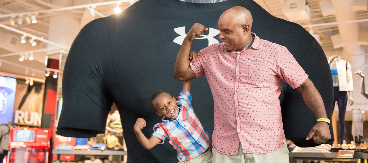 A young boy makes a muscle as he hangs from his dad’s bicep in front of a larger than life Under Armour T shirt
