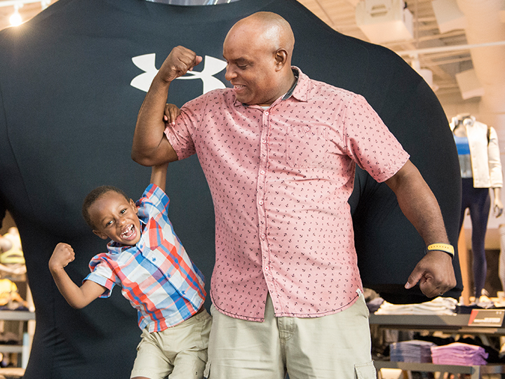 A young boy makes a muscle as he hangs from his dad’s bicep in front of a larger than life Under Armour T shirt