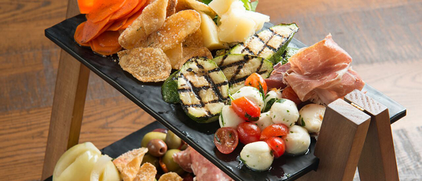 A 2-tiered serving tray containing prosciutto, mozzarella, salami, tomatoes, pickled vegetables, grilled zucchini, arugula, olives and potato chips