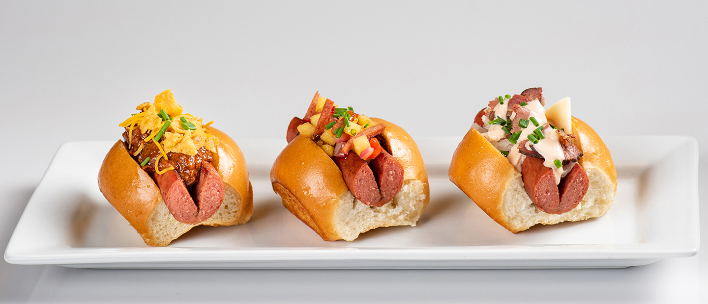 Three specialty mini hot dogs on split top buns, including one topped with chili and cheese, one with diced Spam and pineapple salsa and one with corned beef, Swiss cheese and Russian dressing