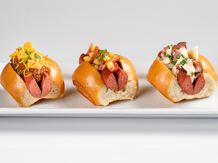 Three specialty mini hot dogs on split top buns, including one topped with chili and cheese, one with diced Spam and pineapple salsa and one with corned beef, Swiss cheese and Russian dressing