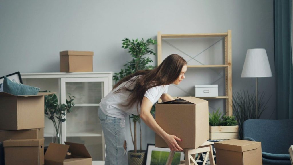 A woman is lifting a box in a room with unpacked items.