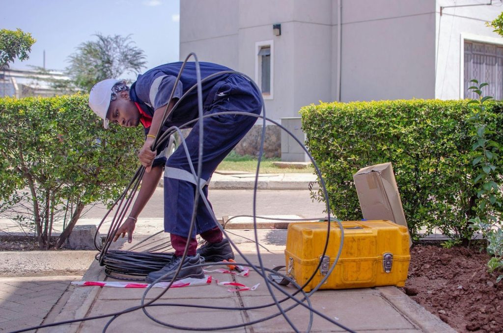 A utility worker is bending down on a sidewalk, organizing and connecting cables beside a toolbox and equipment.