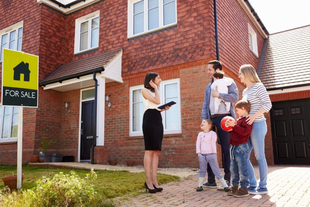 A real estate agent stands outside a brick house with a “For Sale” sign, speaking to a family of four.