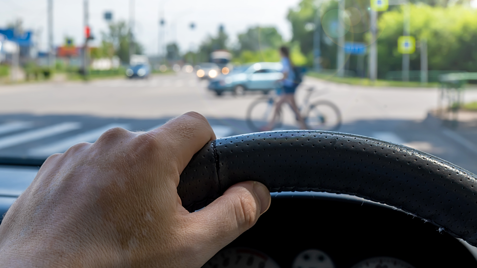 Drivers hand on a steering wheel with a person and bicycle at a crosswalk ahead.