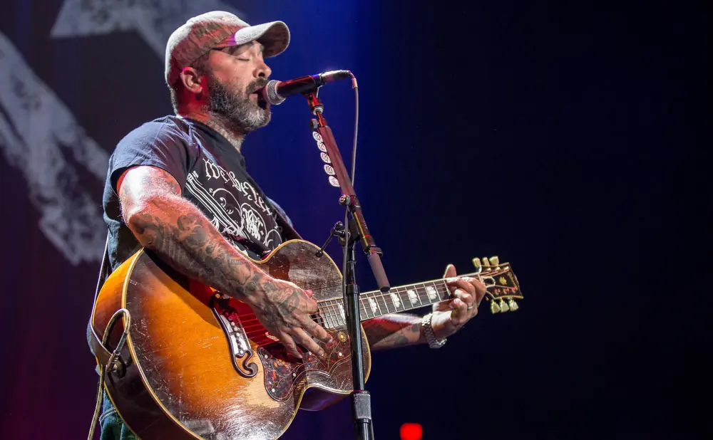 Man wearing a baseball cap and tattoos sings into a microphone while playing an acoustic guitar on stage.