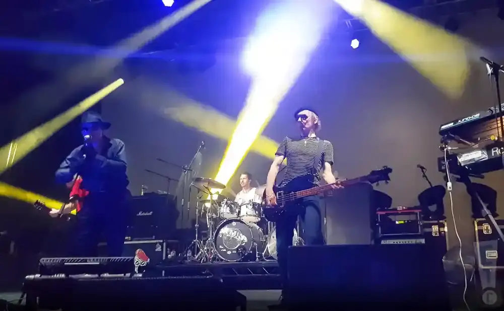 A band performs on a stage with bright blue and yellow lights. A bassist in a striped shirt plays center stage, while a guitarist in a hat sings on the …