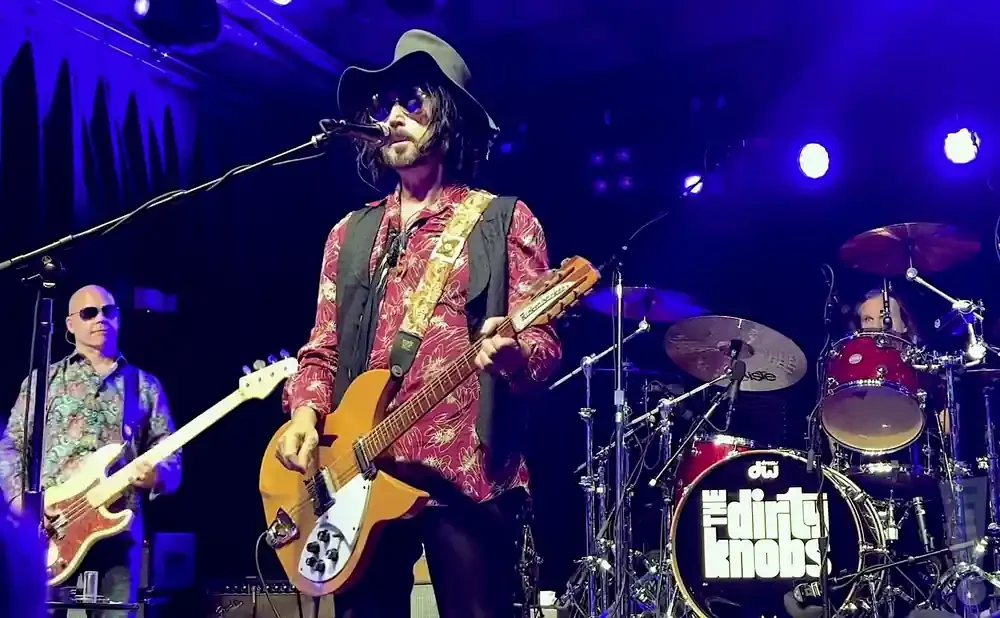 A band plays on stage under blue lights. The lead singer, wearing a hat and sunglasses, sings and plays guitar.